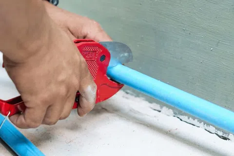 Hands using red pipe cutter to cut blue plastic plumbing pipe near wall.