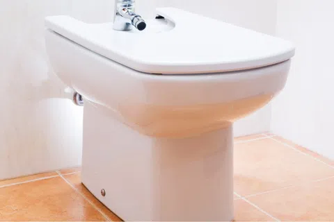 White ceramic bidet with chrome faucet on tan tiled bathroom floor and beige walls
