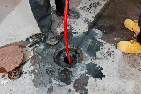 Workers removing blockage from a manhole with a red hose on a worn concrete surface.