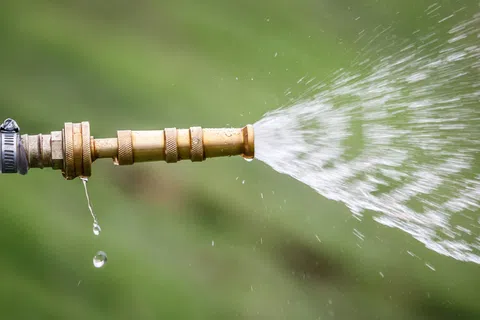 Close-up of a garden hose nozzle spraying water with droplets falling against a blurred green background.