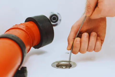 Hand using a drain snake tool to unclog a bathtub drain next to an orange pipe fitting.