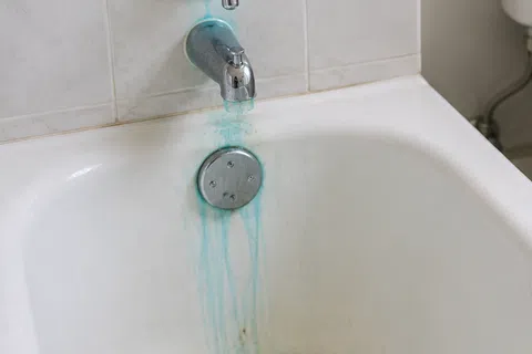 Blue-green stains from running water visible on bathtub, bathroom sink, and toilet bowl in a white tiled bathroom.