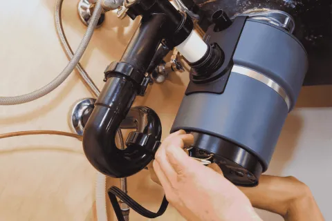 Hands installing a modern kitchen garbage disposal unit under a sink with black plumbing pipes and metal hose connections