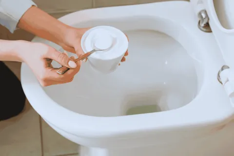 Person holding baking soda cup and spoon over a clean white toilet bowl for cleaning.