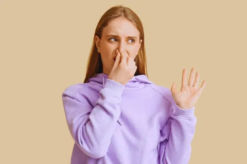 Young woman in purple hoodie holding nose and showing disgust on beige background