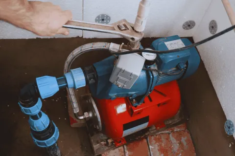 Hand using wrench to tighten pipe fitting on a blue and red water pump system in a utility room.