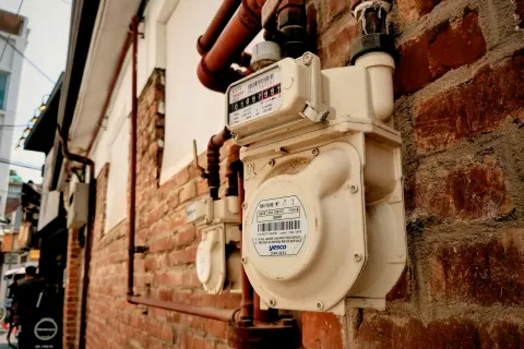 Close-up of a white gas meter mounted on a brick wall with metal pipes and urban background.