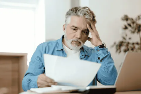 Older man with gray hair and beard looking stressed while reviewing documents at desk with laptop