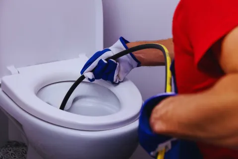 Plumber using a drain snake tool to unclog a white toilet wearing gloves and red shirt.