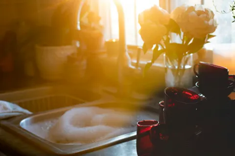 Sunlit kitchen sink filled with soapy water next to stacked red glass cups and a vase of white flowers on the counter