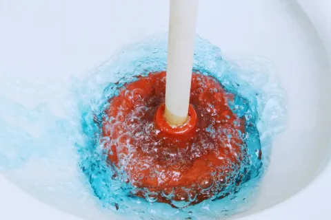 Red plunger creating bubbles as it unclogs blue water in a white sink drain.