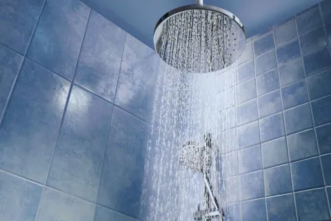Modern chrome rainfall showerhead with water flowing in a tiled blue bathroom.