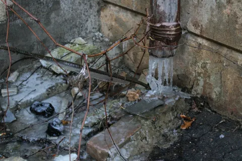 Rusty pipe with frozen water and icicles against aged concrete wall outdoors in winter.