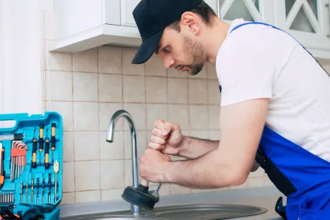 Plumber in blue overalls using a plunger on a kitchen sink faucet with tools nearby on the counter.
