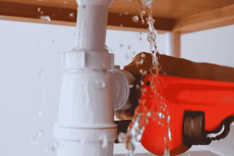 Close-up of a leaking pipe being tightened with a red pipe wrench under a wooden sink cabinet.