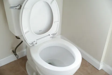 Clean white toilet with open lid in a beige tiled bathroom corner near white walls and baseboard.