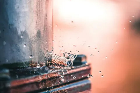 Water droplet splashing on rusty metal pipe with blurred warm sunlight background.