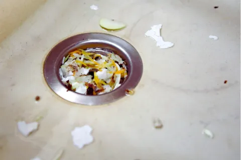 Close-up of a kitchen sink drain clogged with food scraps and vegetable peels on a beige surface.