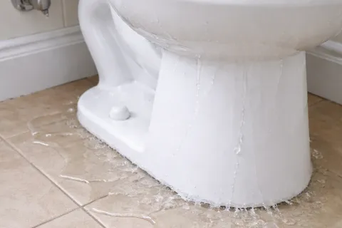 White toilet leaking water onto beige tiled bathroom floor causing a plumbing issue.