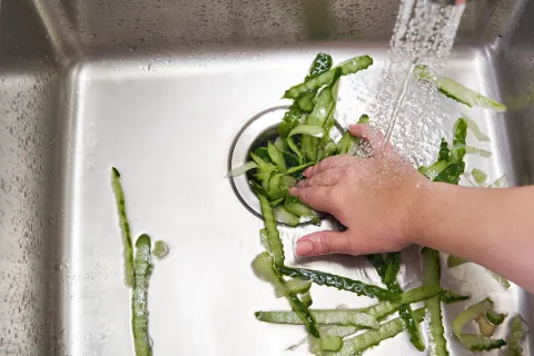 Hand washing green vegetable peels under running water in a stainless steel kitchen sink drain.