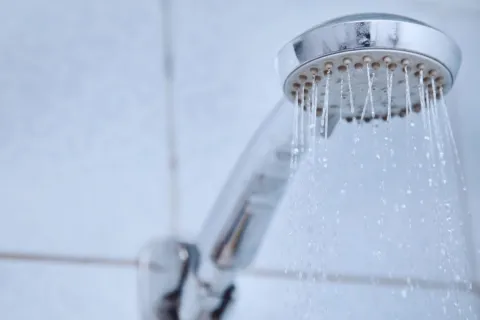 Close-up of a chrome showerhead with water droplets flowing against a tiled bathroom wall.