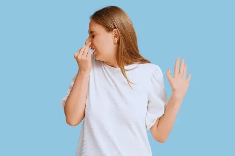 Young woman in white shirt holding her nose with a disgusted expression on a blue background