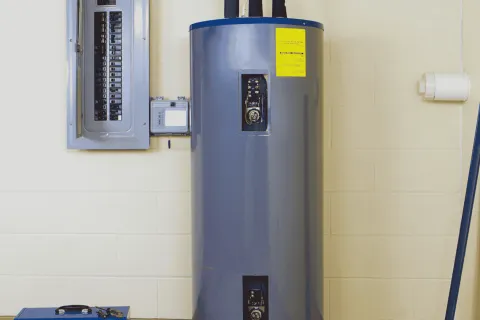 Large gray electric water heater next to a circuit breaker panel mounted on a beige wall in a utility room.
