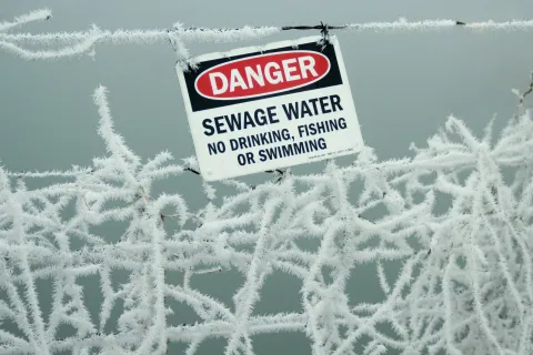 Danger sign warning of sewage water, no drinking, fishing, or swimming, on frost-covered wire fence.