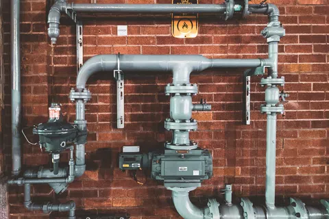 Industrial gas pipeline system with valves and meters mounted on a red brick wall with a gas hazard sign.