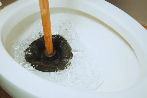 Close-up of a toilet bowl being unclogged with a wooden-handled plunger creating water pressure.