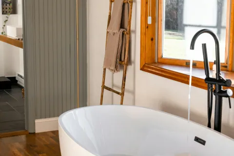 Modern freestanding white bathtub with running faucet, wooden ladder towel rack, and large window with natural light.