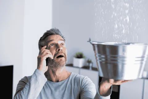 Man on phone holding a metal bucket catching water leaking from ceiling inside a home.