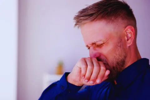 Man in blue shirt covering mouth and nose, showing signs of sneezing or illness indoors.