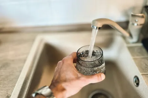 Hand filling a glass with water from a kitchen faucet over a beige sink with metal drain.