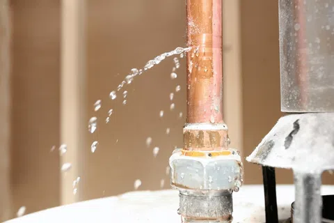 Water leaking from a hole in a copper pipe connected to a metal valve inside a residential area.