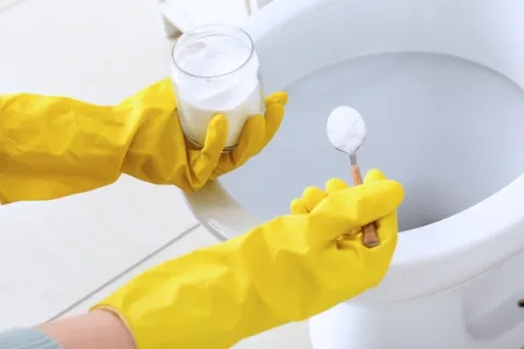 Person wearing yellow gloves holding baking soda to clean a white toilet bowl in a bathroom.
