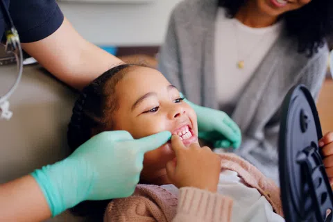 Child smiling while dentist with gloves examines teeth, mother holding mirror in dental clinic.