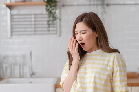 Young woman in striped shirt covering her mouth while yawning in a bright kitchen setting