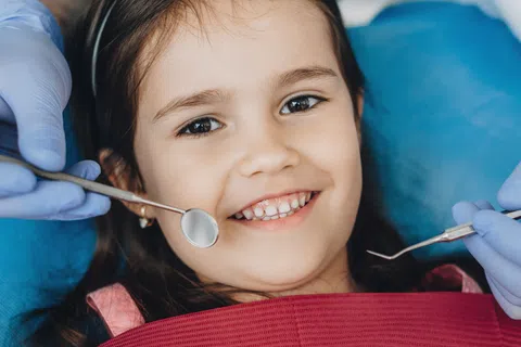 Smiling young girl sitting in dental chair with dentist's hands holding dental tools near her mouth