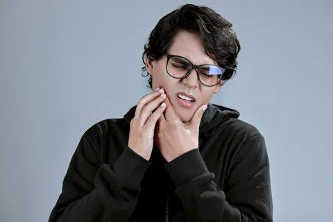 Young man in black hoodie wearing glasses holding his jaw in pain against a plain gray background