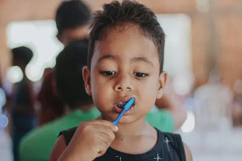 Young boy brushing his teeth with a blue toothbrush in a busy indoor setting.