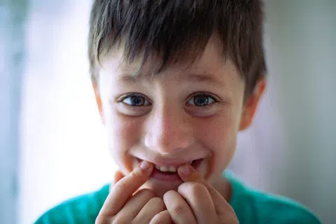 Close-up of a smiling boy with blue eyes and freckles showing his teeth with fingers indoors