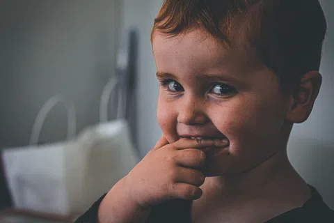 Close-up of a smiling young boy with brown hair, fingers near mouth, looking playfully at the camera indoors.