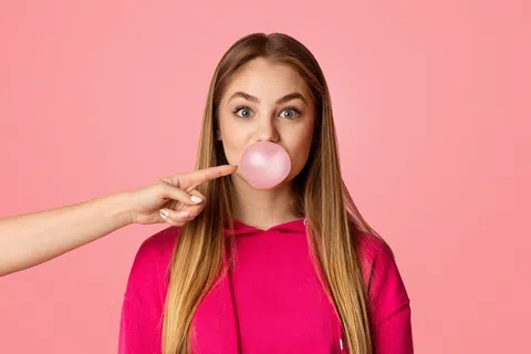 Young woman blowing a pink bubblegum bubble with a finger about to pop it against a pink background.