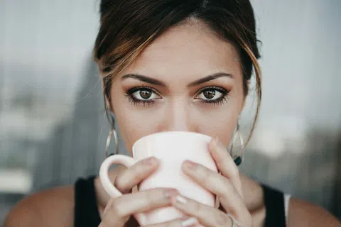 Close-up of a woman with striking eyes holding and sipping from a white mug outdoors.