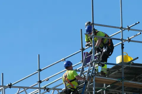 Two construction workers in safety gear working on metal scaffolding under clear blue sky.