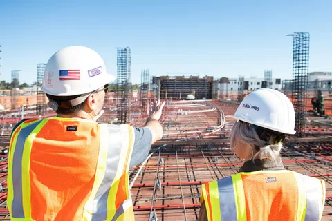 Two construction workers in safety gear inspecting a large concrete rebar framework at a building site under clear sky.