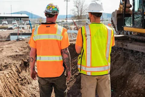 Two construction workers in safety vests and helmets overseeing excavation site with heavy machinery in background.