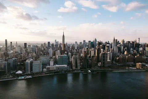 Aerial view of New York City skyline with skyscrapers along the waterfront under a partly cloudy sky.