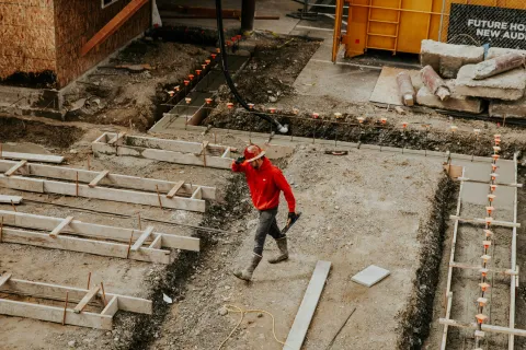 Construction worker in red jacket and helmet walking on foundation site with wooden frames and dirt trench